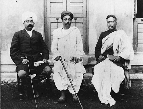 Photograph of three seated men in front of a shuttered building: Lala Lajpat Rai in a turban and dark coat on the left, Bal Gangadhar Tilak in the centre in a turban and white dhoti holding a walking stick, and Bipin Chandra Pal on the right in spectacles with a shawl over his shoulder.