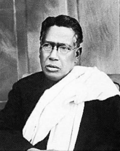 Seated half-length photographic portrait of Bipin Chandra Pal wearing round spectacles, a dark garment and a white shawl draped over one shoulder, looking towards the camera.