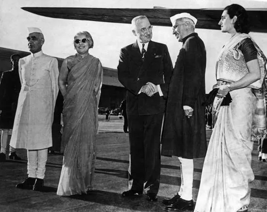 Black and white photograph on an airport apron: a group of five people beside an aircraft, two men shaking hands in the centre, with a woman in a sari and dark glasses and a man in a light sherwani and cap to the left, and a woman in a sari to the right.